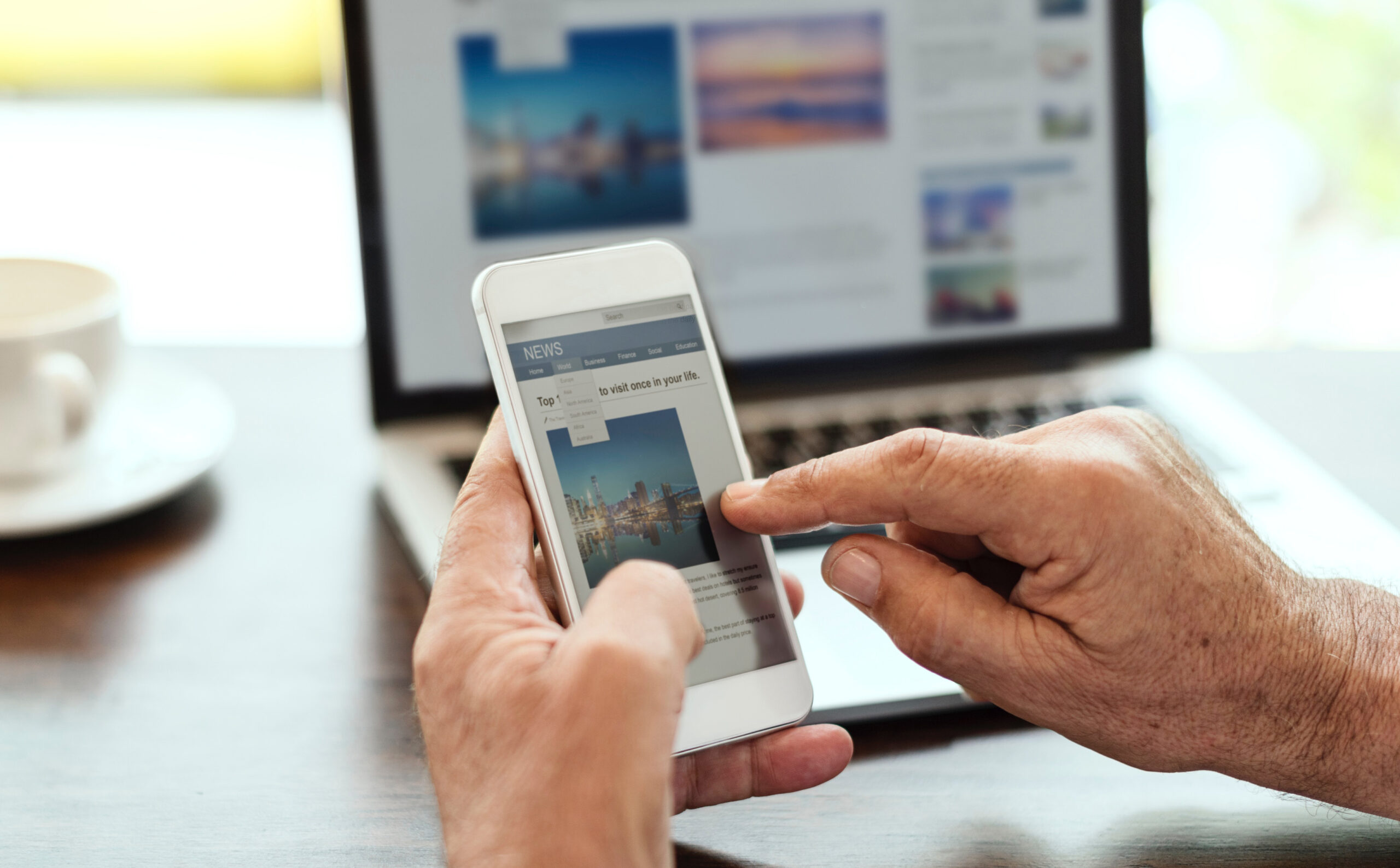 Someone scrolls news on their phone in front of their laptop