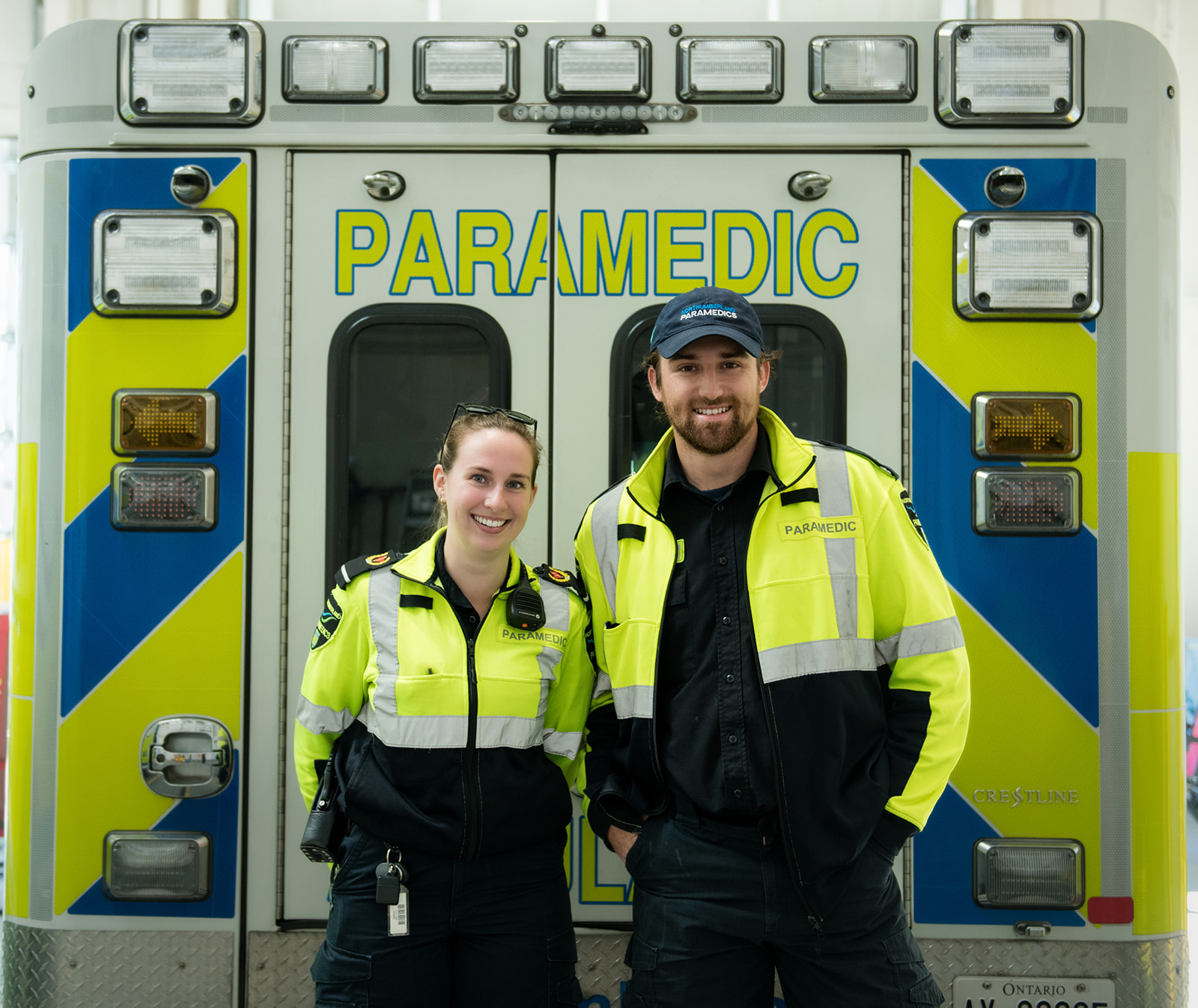Two paramedics smile as they stand in front of the back doors of an ambulance