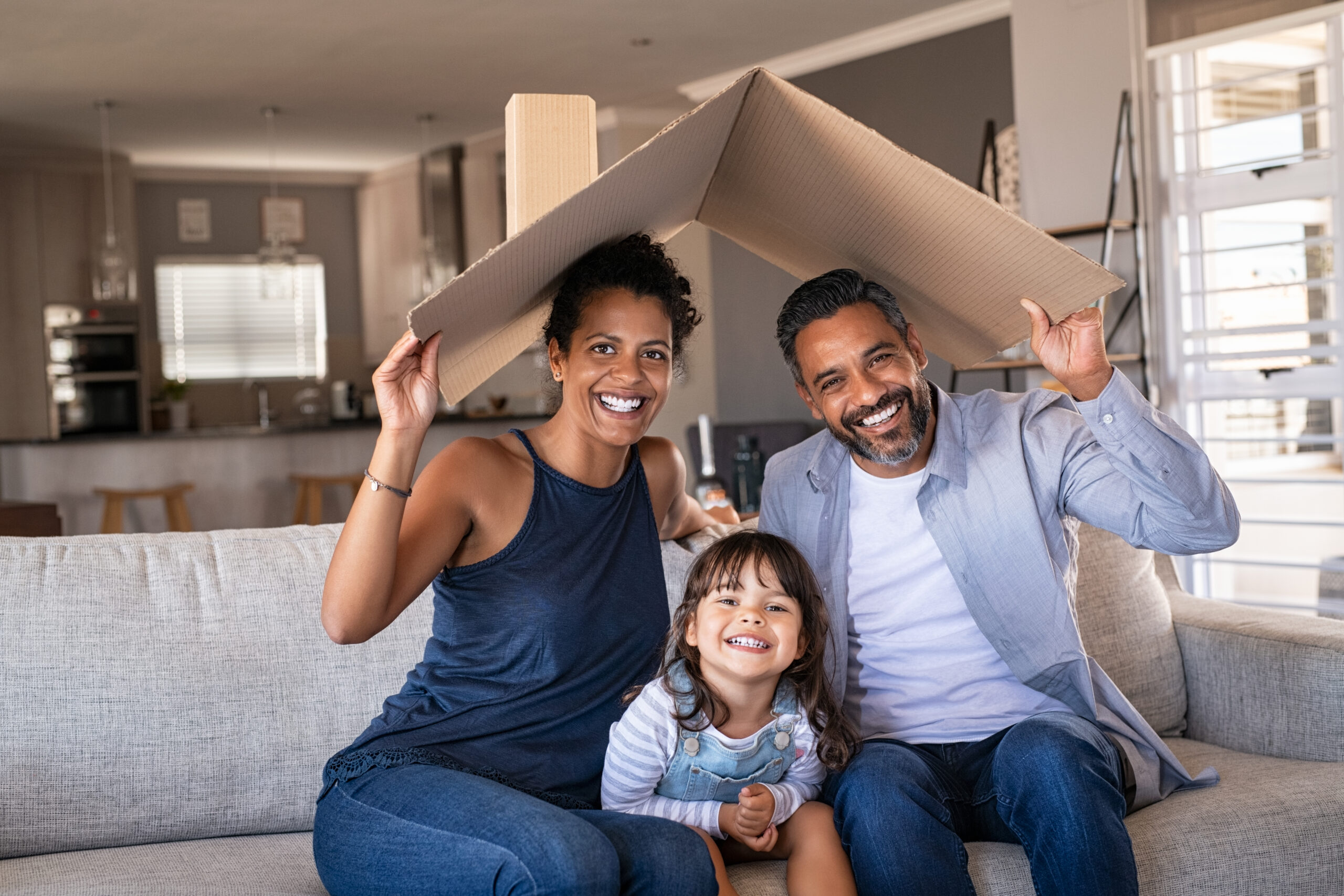 Portrait of smiling family sitting on couch holding cardboard roof over their head.