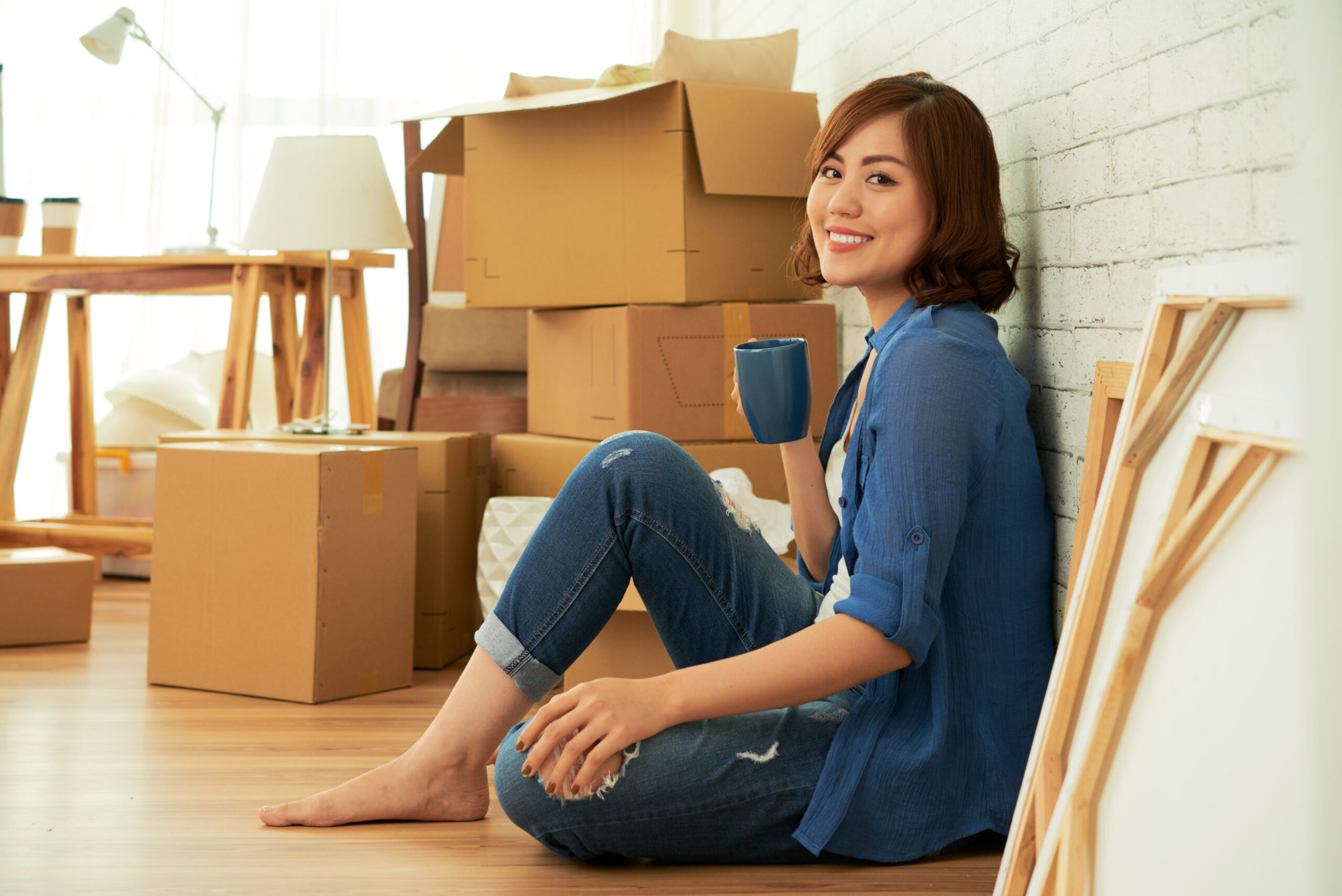 Woman sits on floor of new home with cardboard boxes behind her, drinking coffee